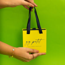 Load image into Gallery viewer, Yellow gift bag with black handles held by a person against a green background